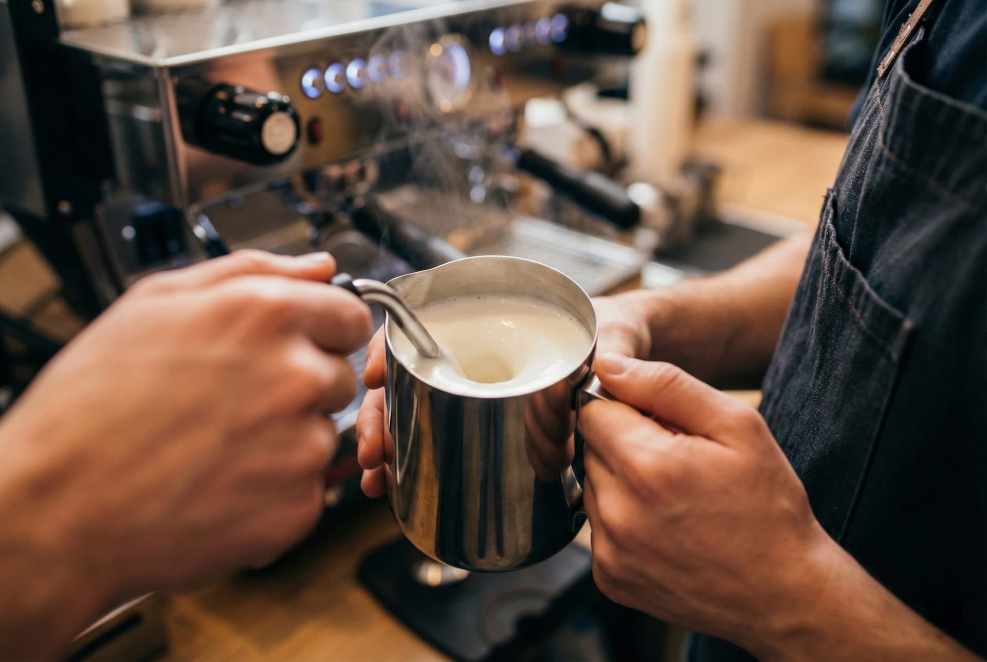 Professional barista demonstrating proper milk steaming technique creating velvety microfoam for flat white using steam wand position and temperature control methods from 15 years training experience