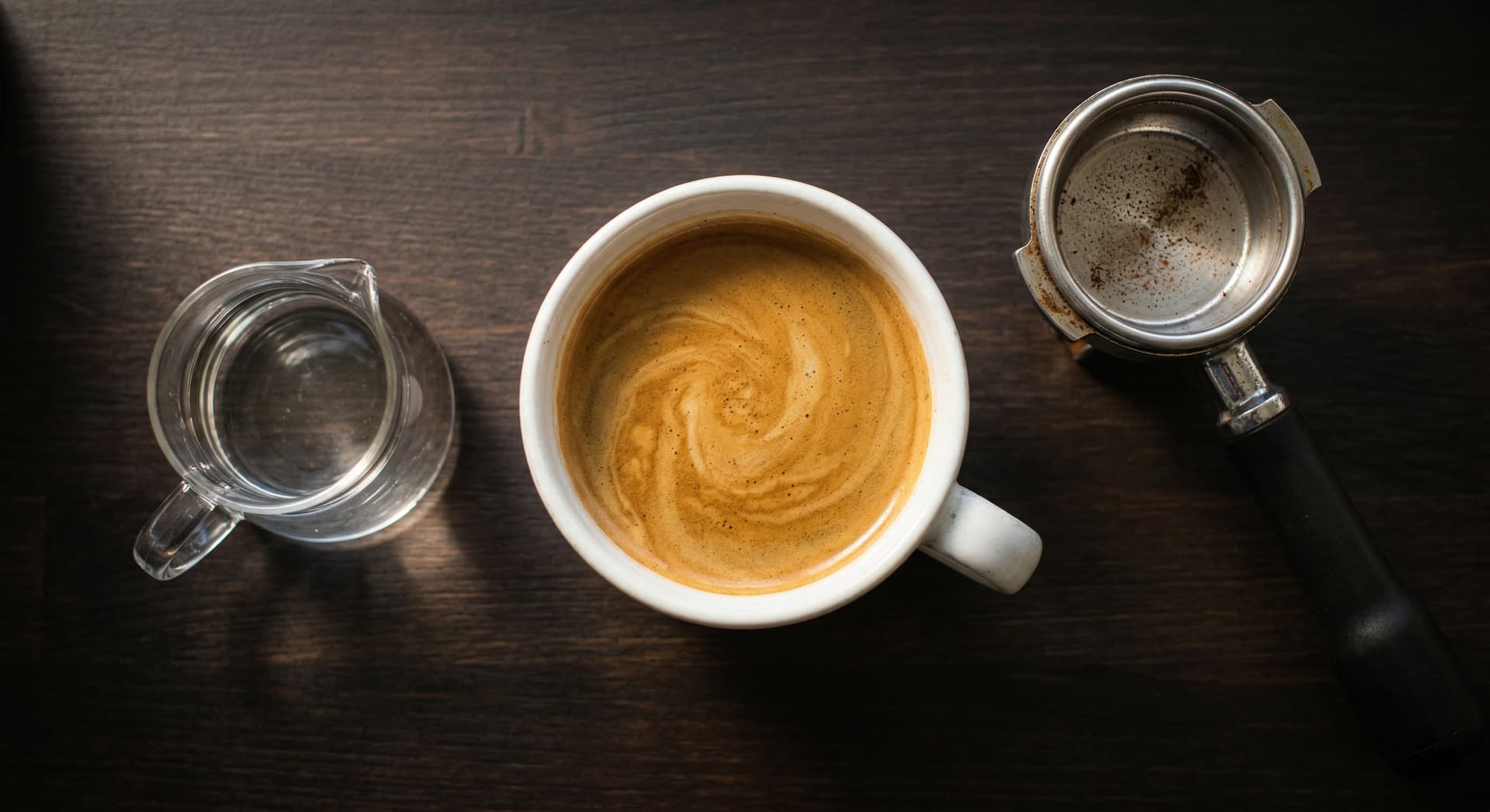 Freshly made café americano in a wide-rimmed white ceramic mug with intact golden crema on a dark espresso-stained wooden café counter