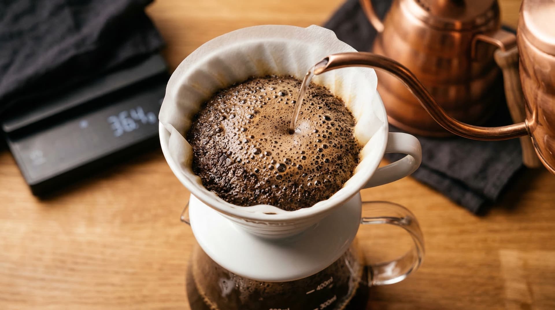 A stainless steel Bialetti Moka pot on a gas burner at medium-low heat with coffee streaming into the upper chamber — stovetop espresso brewing in progress