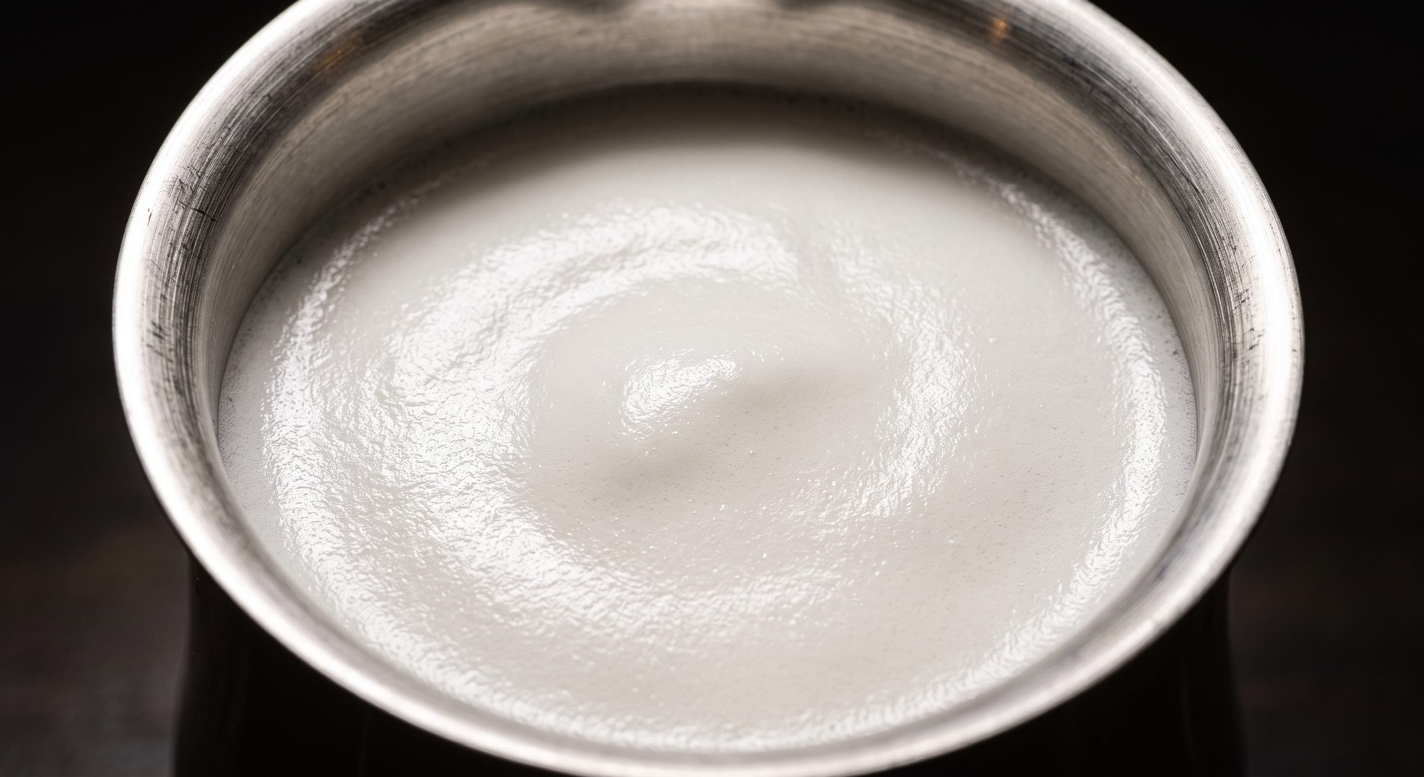 Close-up of correctly textured cappuccino foam in a stainless steel pitcher: thick, dense, glossy white foam with no large bubbles