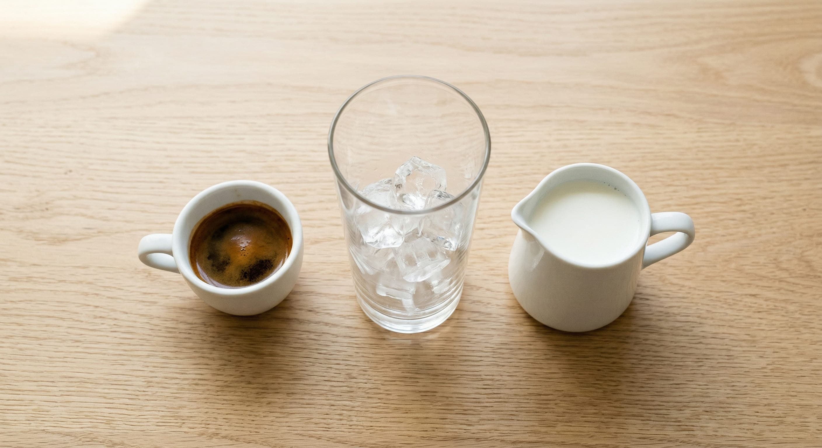 Cup of espresso, a glass of ice and a small jug of milk on a light wooden table