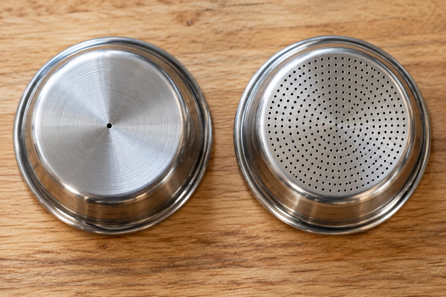 Underside of a dual-wall espresso basket with one small hole next to a single-wall basket with many holes