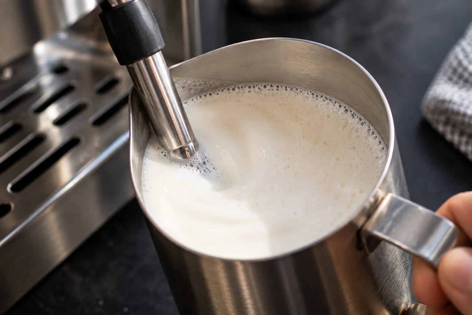 Steam wand tip just below the surface of milk in a stainless steel pitcher, creating a smooth whirlpool