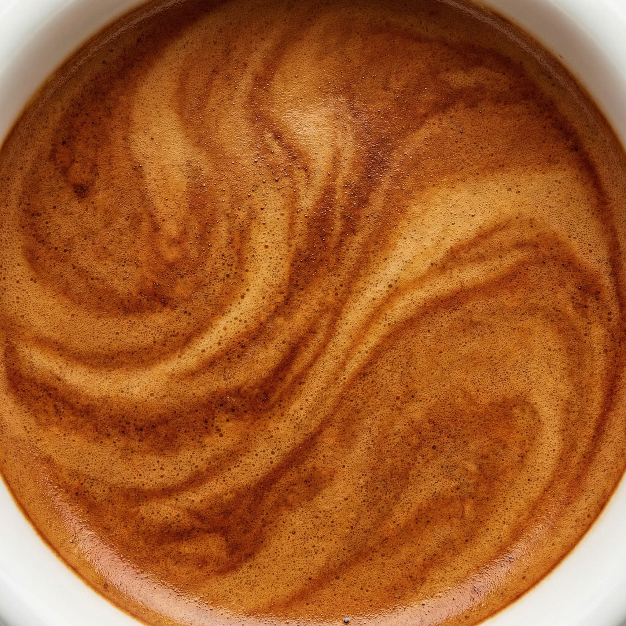 Macro photography of espresso crema showing tiger-stripe pattern, reddish-brown color, fine bubble structure, and persistent foam layer on freshly extracted espresso shot in white ceramic demitasse—ideal crema characteristics