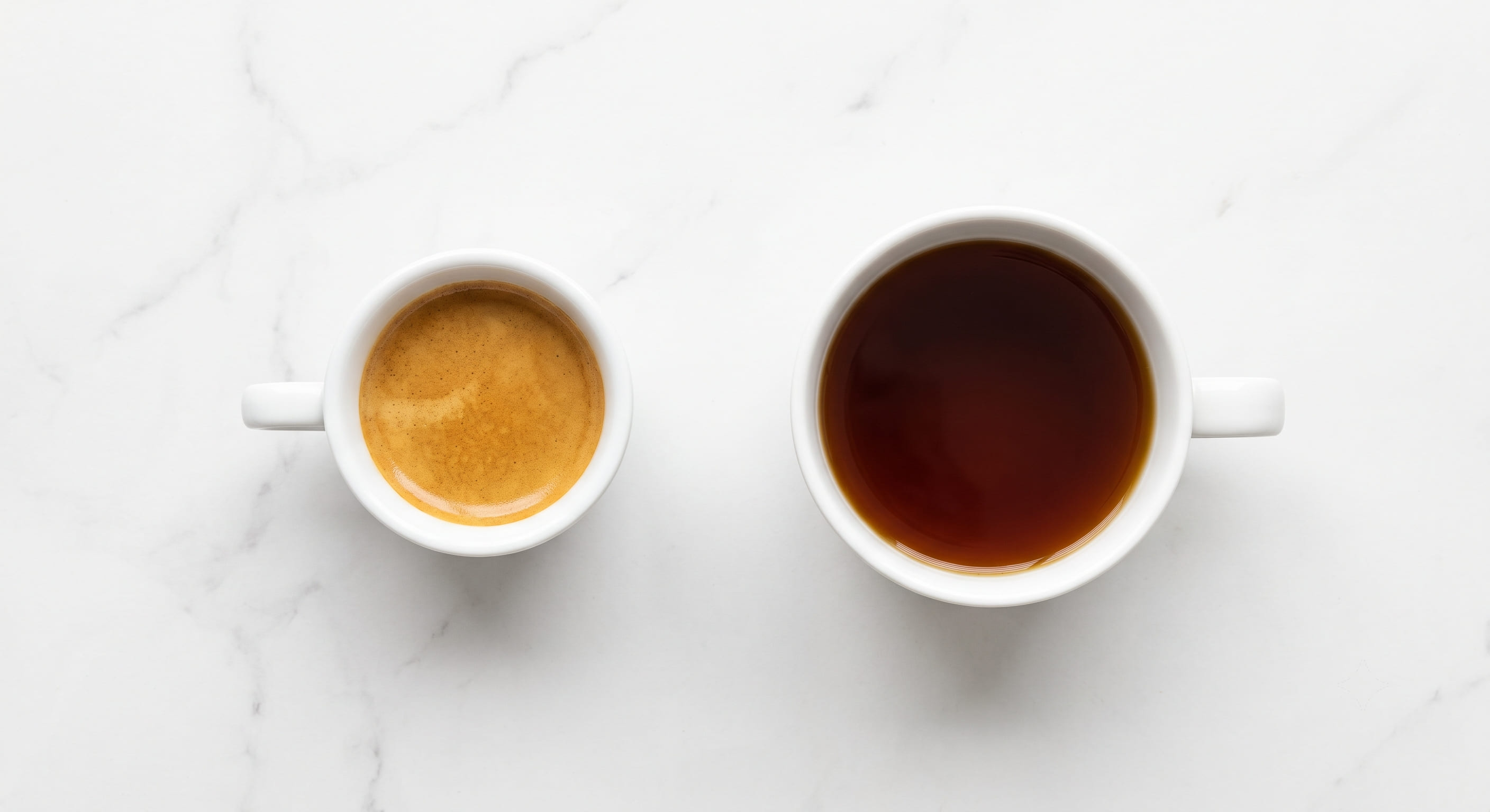 Side-by-side comparison of espresso and drip coffee — small demitasse with golden crema on left, large white ceramic mug of filter coffee on right, on a white marble surface