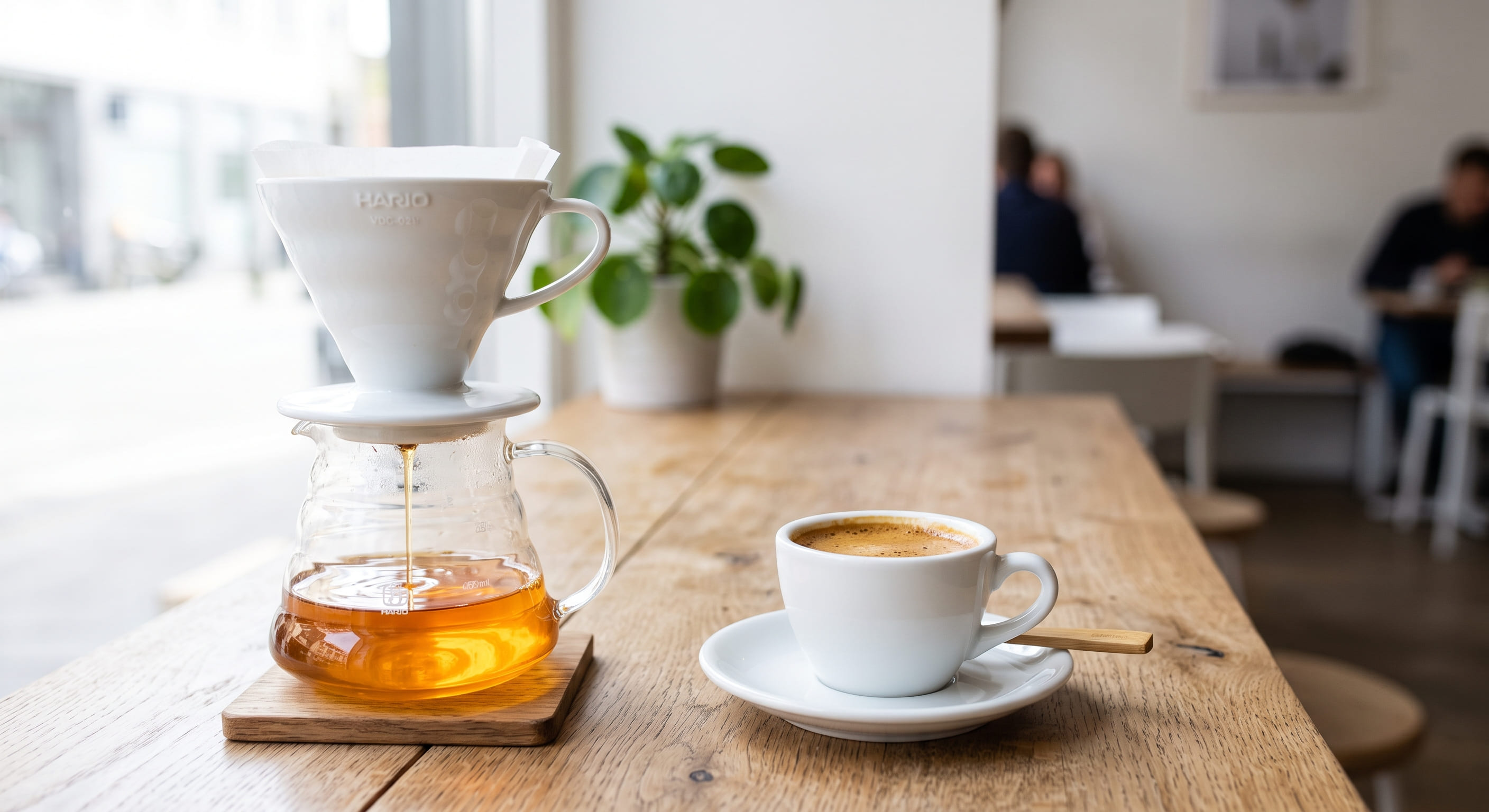 Pour-over coffee dripping through a Hario V60 dripper into a clear glass carafe next to a finished espresso shot in a demitasse — contrasting the clarity and colour of each brewing method