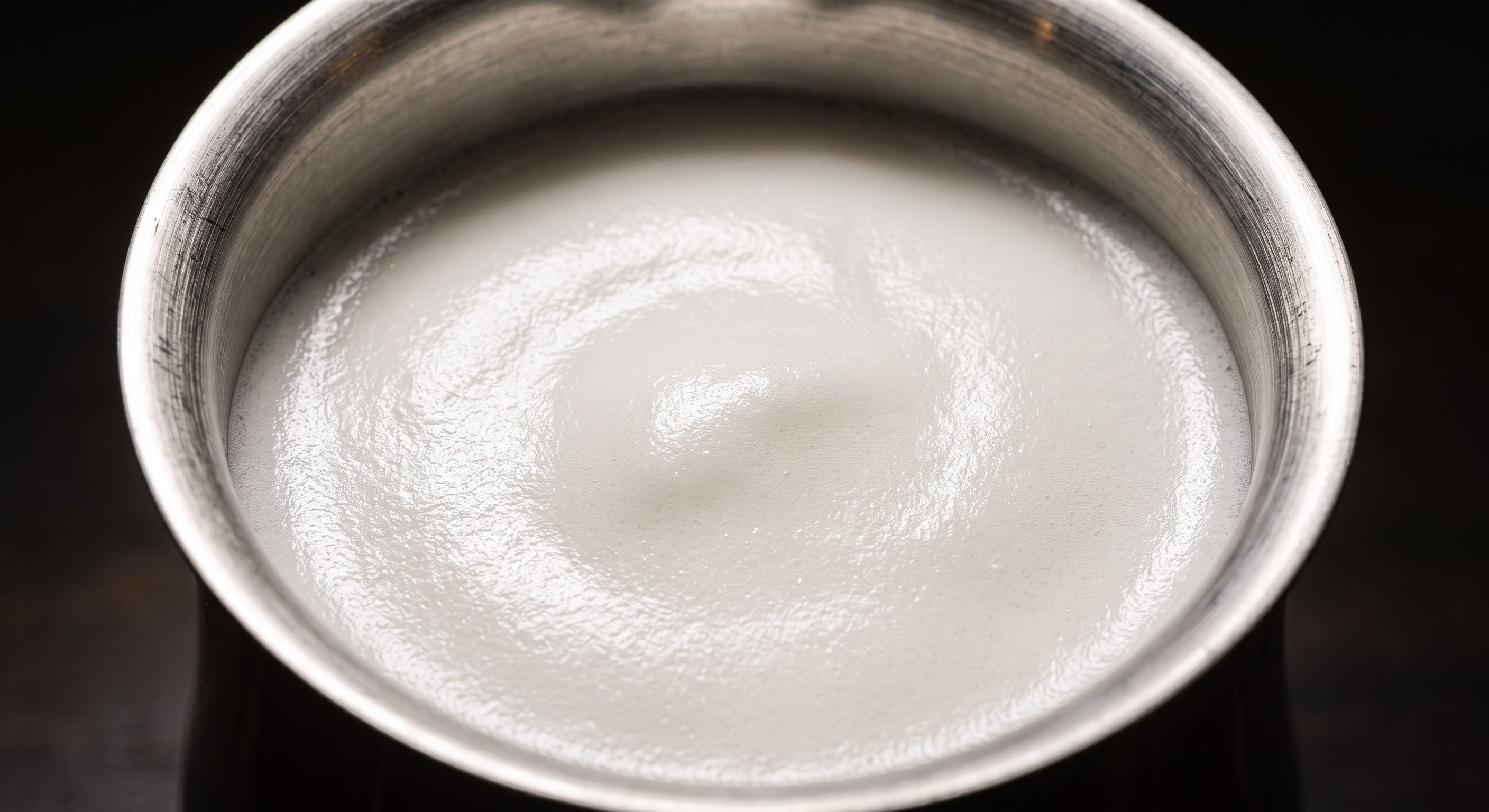 Close-up of correctly textured cappuccino milk foam in a stainless steel pitcher — thick, dense, glossy white foam with no visible large bubbles, showing the correct texture for a traditional cappuccino foam cap