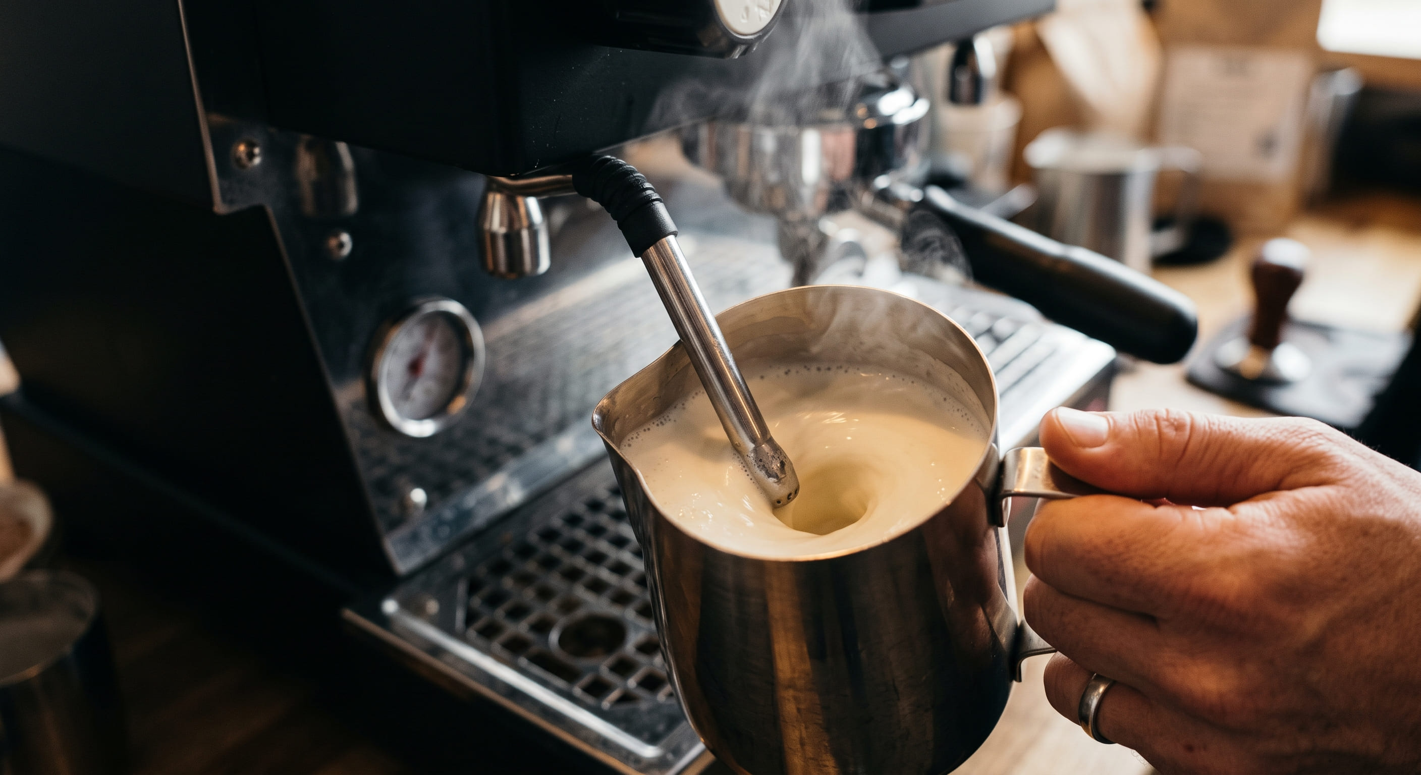 Barista steaming milk for a latte in a stainless steel pitcher with an espresso machine steam wand — showing correct tip position and vortex milk texture technique