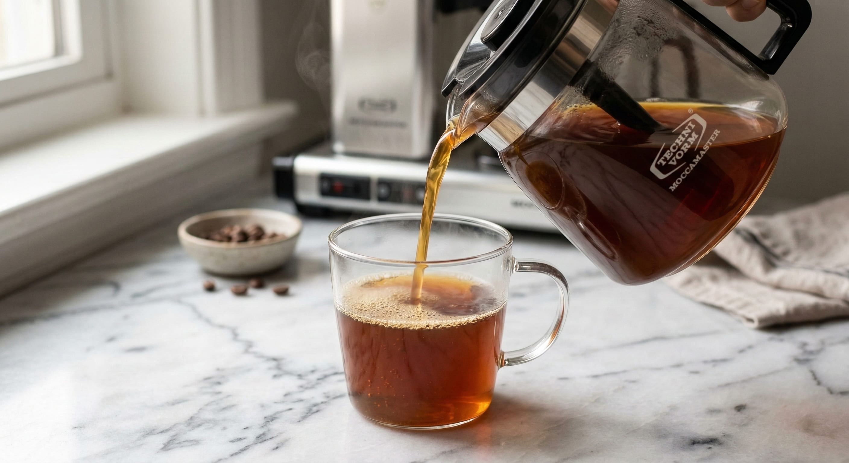 Pouring freshly brewed specialty coffee from Technivorm Moccamaster glass carafe — golden crema visible on surface indicating proper SCA Gold Cup extraction temperature